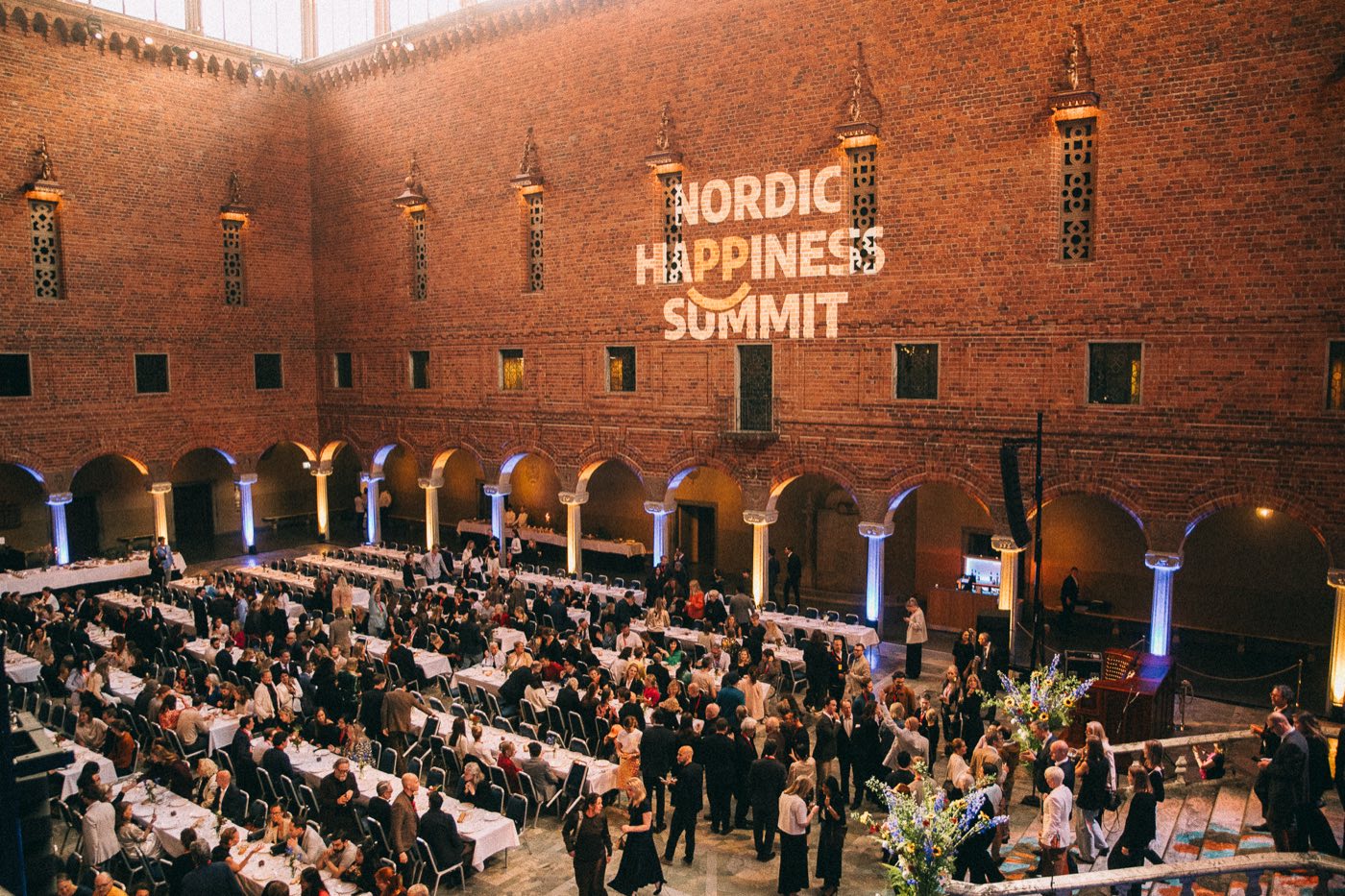 The Blue Hall of Stockholm City Hall filled with guests at long banquet tables, with the Nordic Happiness Summit logo projected on the brick wall