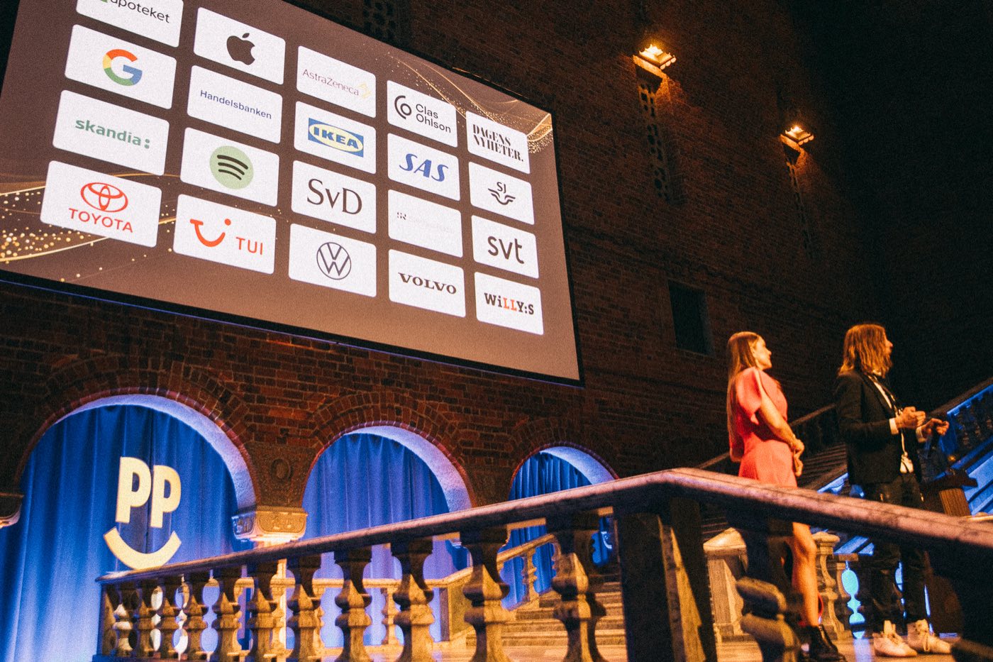 Presenters on the staircase of the Blue Hall next to a large screen showing the organizations included in the first Happiness Impact study